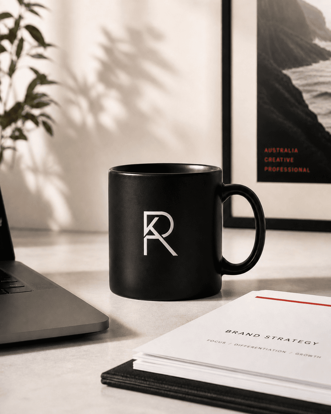 A black ceramic mug stamped with the RAKA monogram, sitting on a clean studio desk next to a Brand Strategy booklet